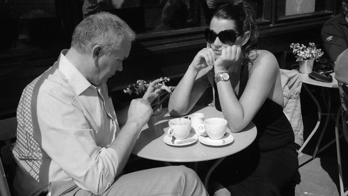 Cặp đôi ngồi đối diện trong quán cà phê với một người dán mắt vào điện thoại minh họa hành vi phubbing gây xói mòn gắn kết